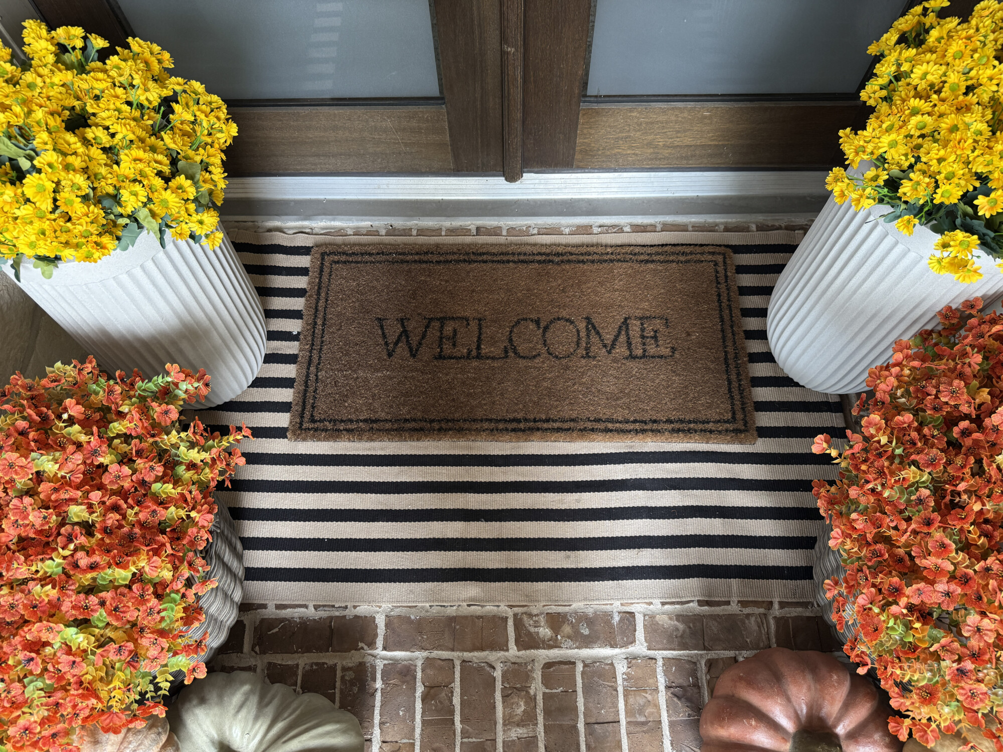 Fall front porch decorating with layered striped doormat, welcome mat, white planters filled with yellow mums and orange flowers, and neutral pumpkins for cozy autumn front door styling ideas.