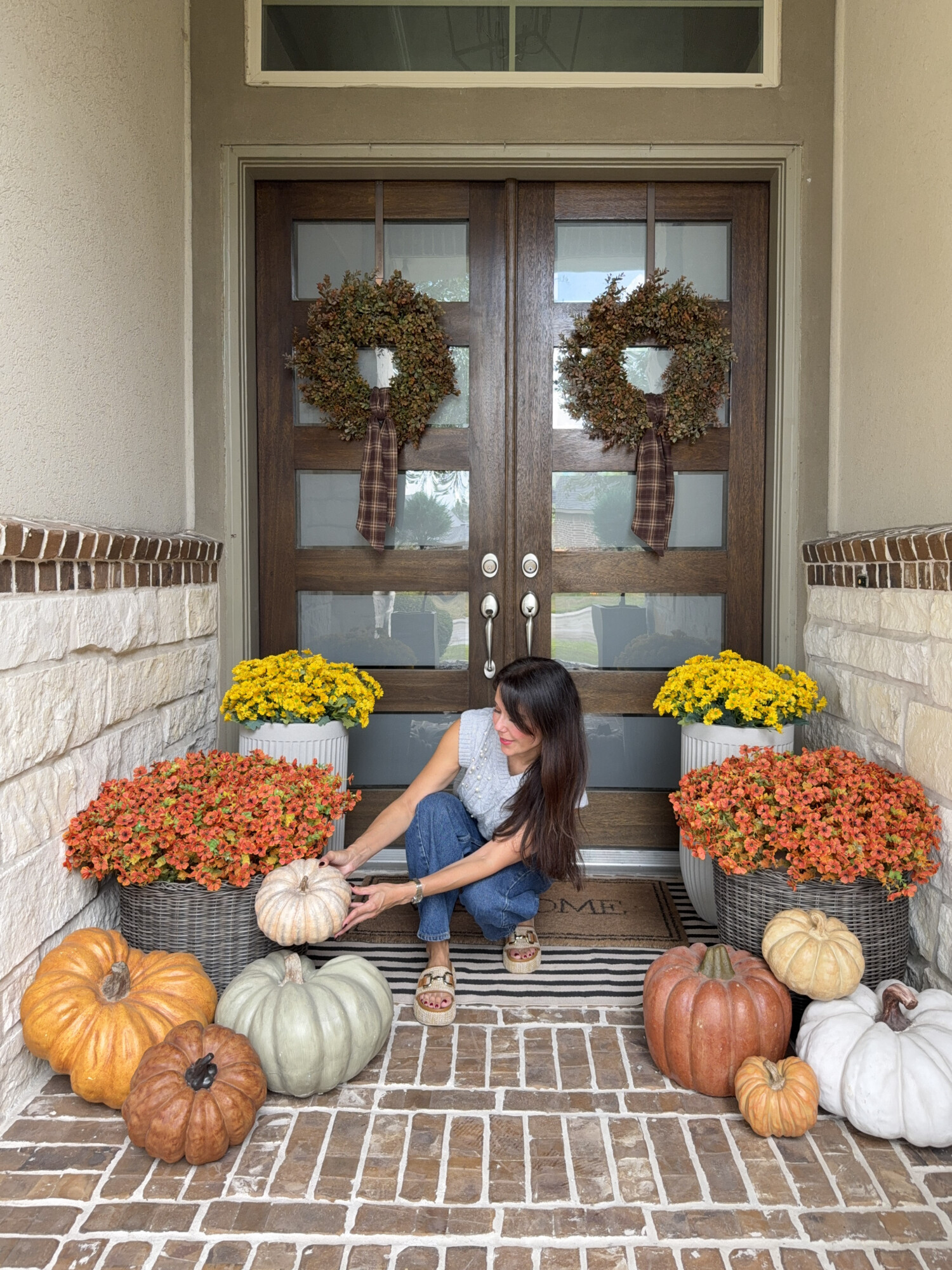 Fall front porch decorating with layered doormats, pumpkins in orange, green, and white tones, yellow mums, and orange flowers in planters. Woman arranging pumpkins on autumn porch with double wooden doors and matching fall wreaths.