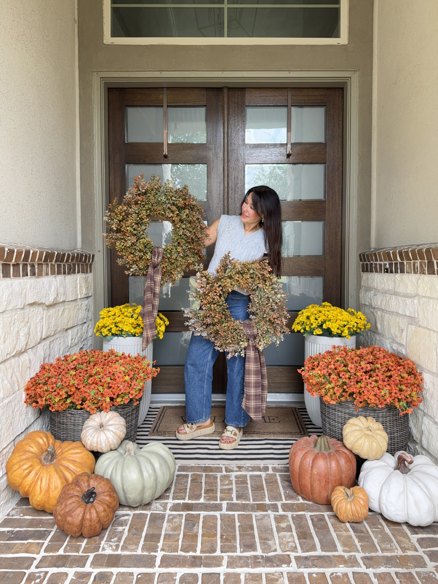 Fall front porch styling with pumpkins in orange, green, and white, colorful mums in planters, and woman holding two autumn wreaths with plaid ribbon in front of double wooden doors for cozy fall entryway decorating ideas.