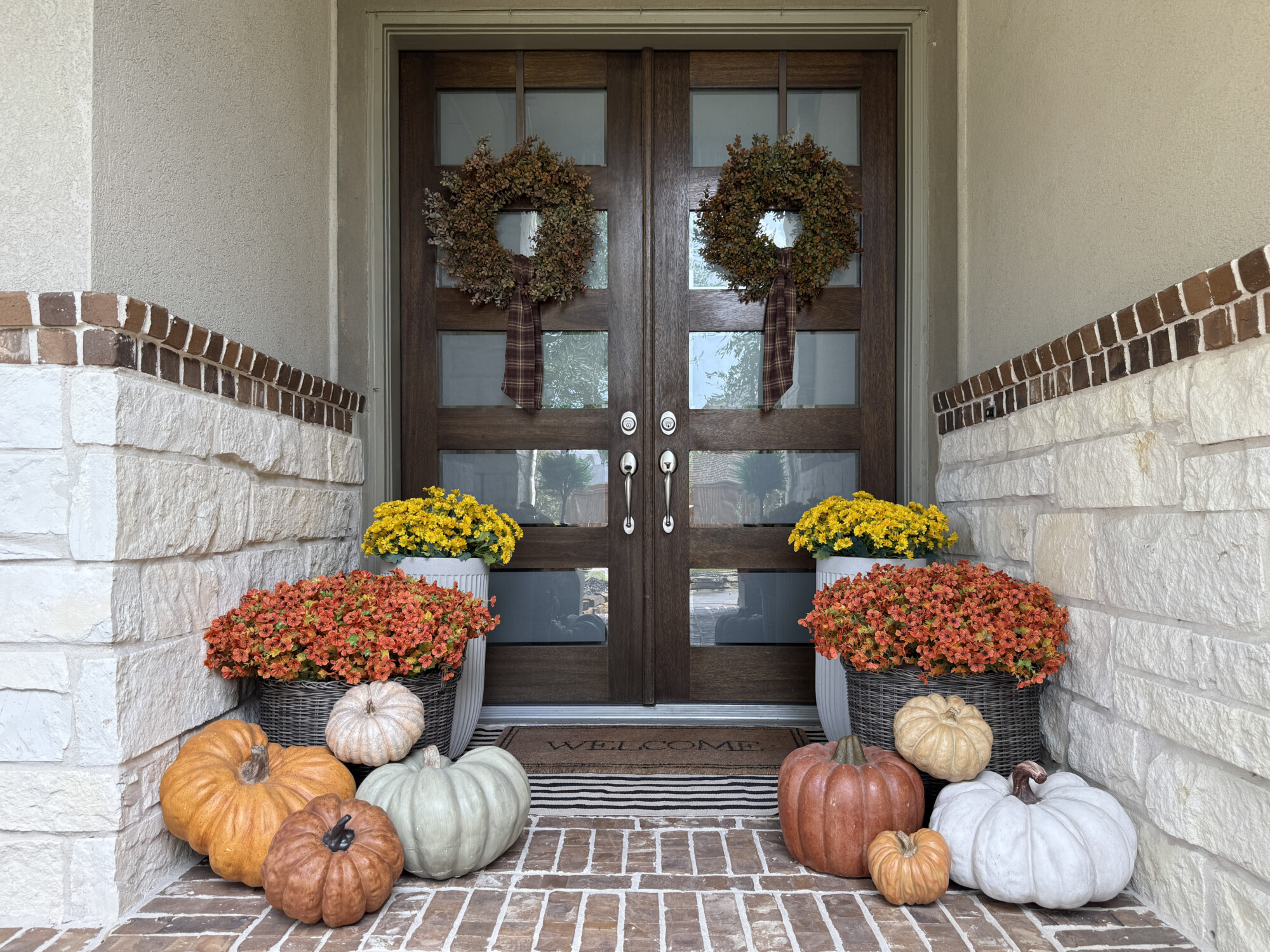 Fall front porch with double wooden doors, matching autumn wreaths, yellow mums, orange flowers in white and gray planters, layered welcome doormat, and pumpkins in neutral and classic fall colors for cozy front door decorating ideas.