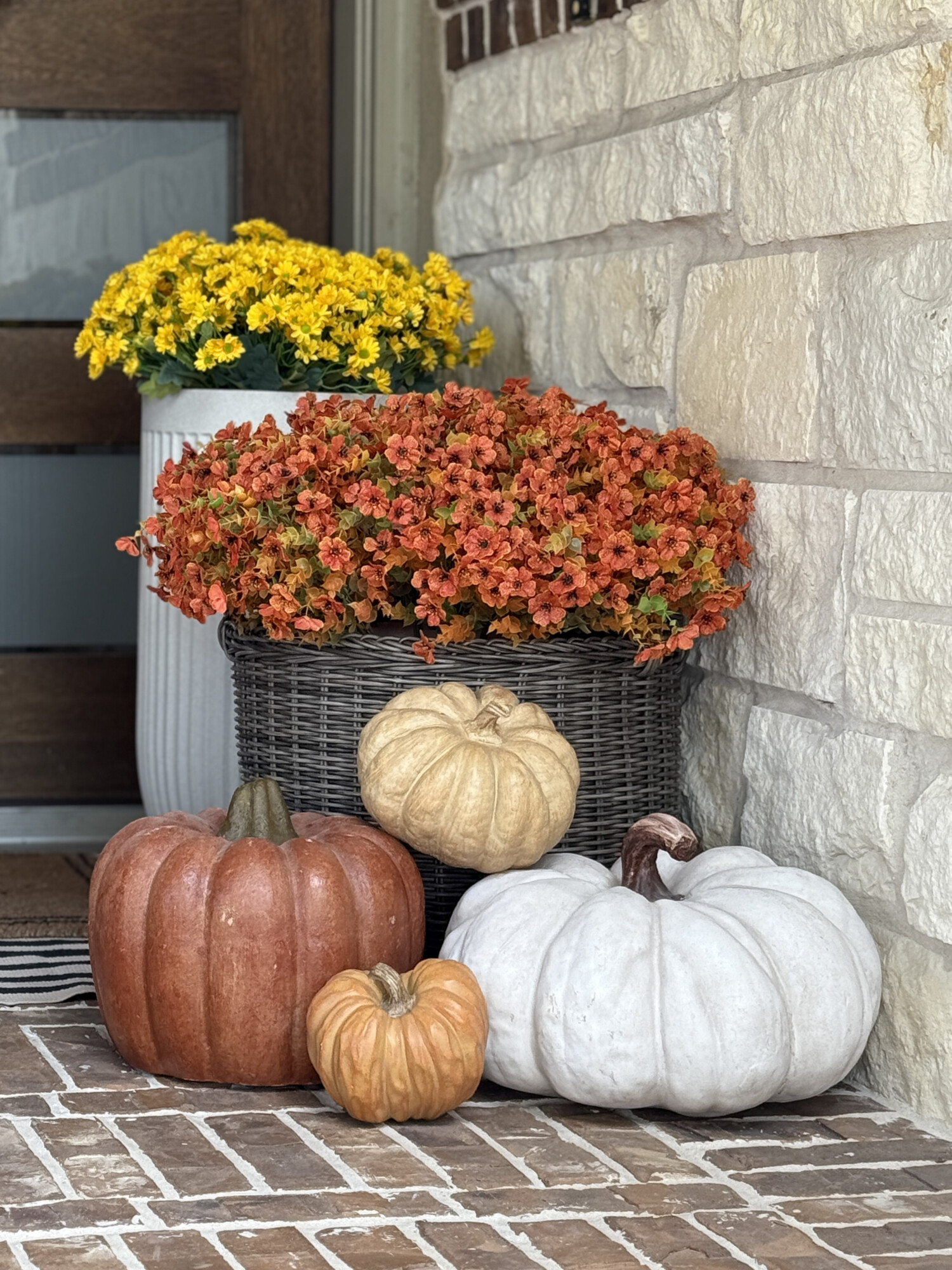 Fall porch detail with orange, white, and neutral pumpkins arranged on brick steps, wicker planter filled with orange flowers, and white planter with yellow mums for seasonal autumn front door decorating ideas.