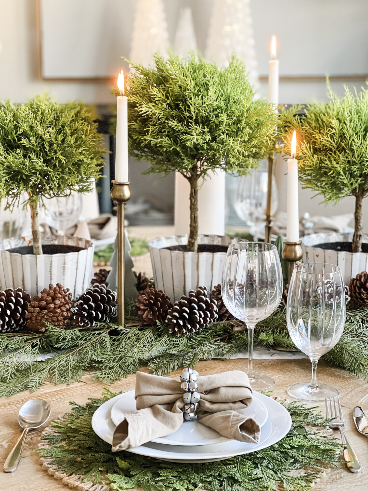 Elegant holiday table setting with fresh greenery, pinecones, and topiary trees in white pots surrounded by glowing candles and neutral dinnerware, creating a cozy and sophisticated Christmas tablescape.