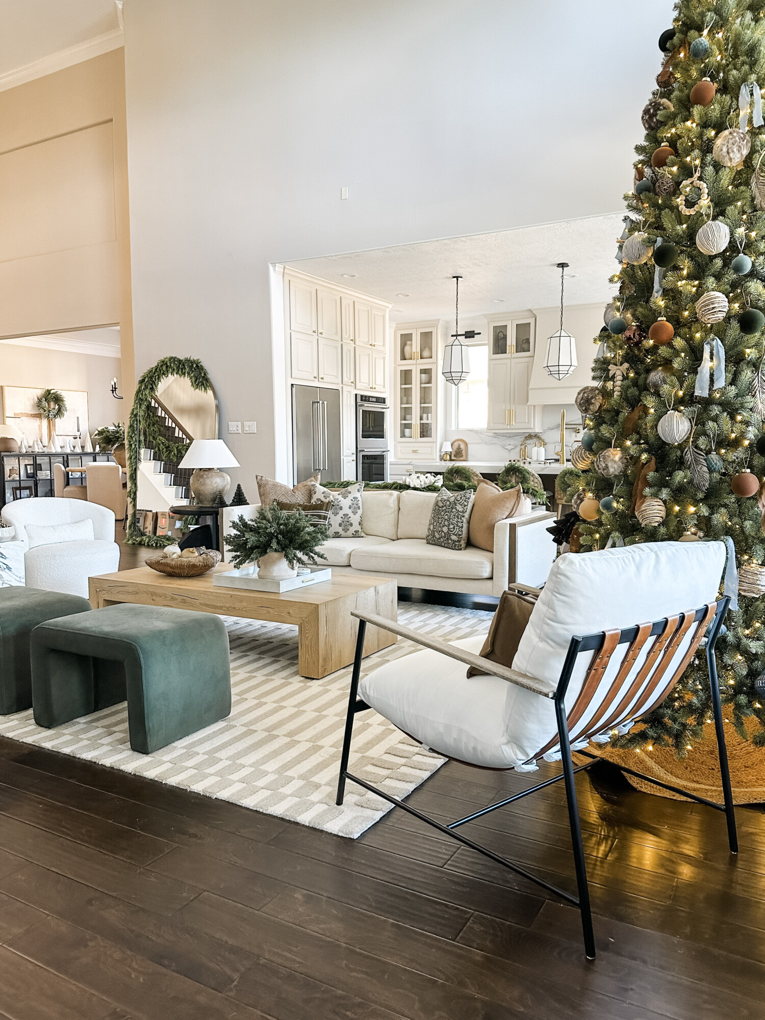 Cozy living room with tall Christmas tree, neutral sofa, green velvet stools, and holiday greenery décor