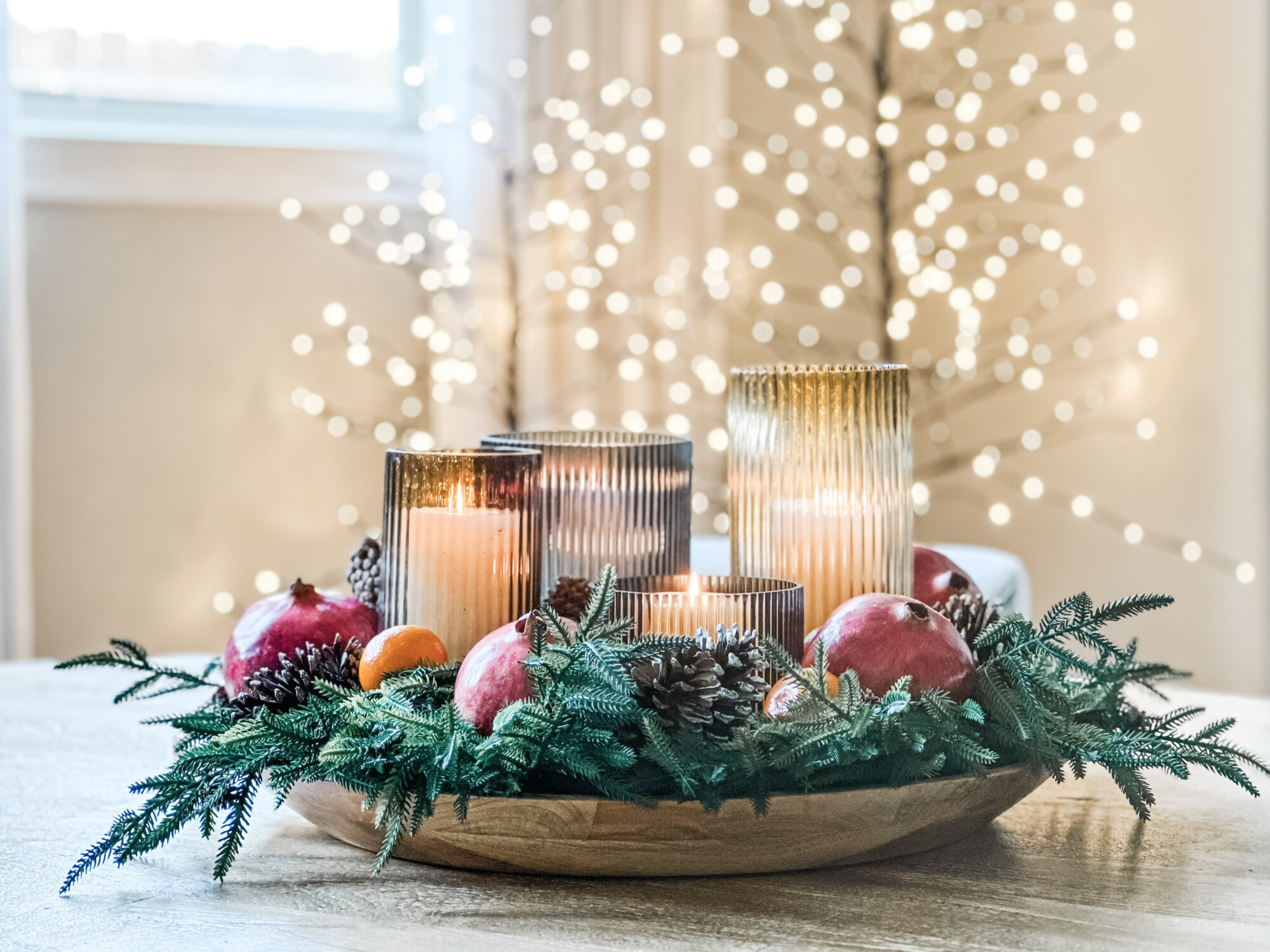 Festive Christmas centerpiece featuring ribbed glass candles, pinecones, pomegranates, and evergreen branches arranged in a wood bowl for a warm, natural holiday table look.
