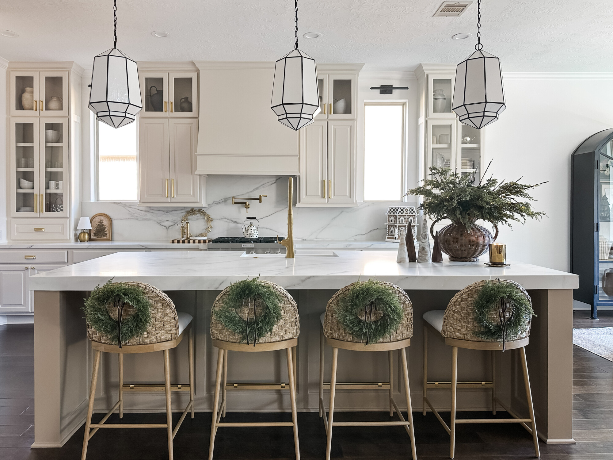 Neutral holiday kitchen with marble island, woven barstools decorated with greenery wreaths, and natural Christmas décor accents
