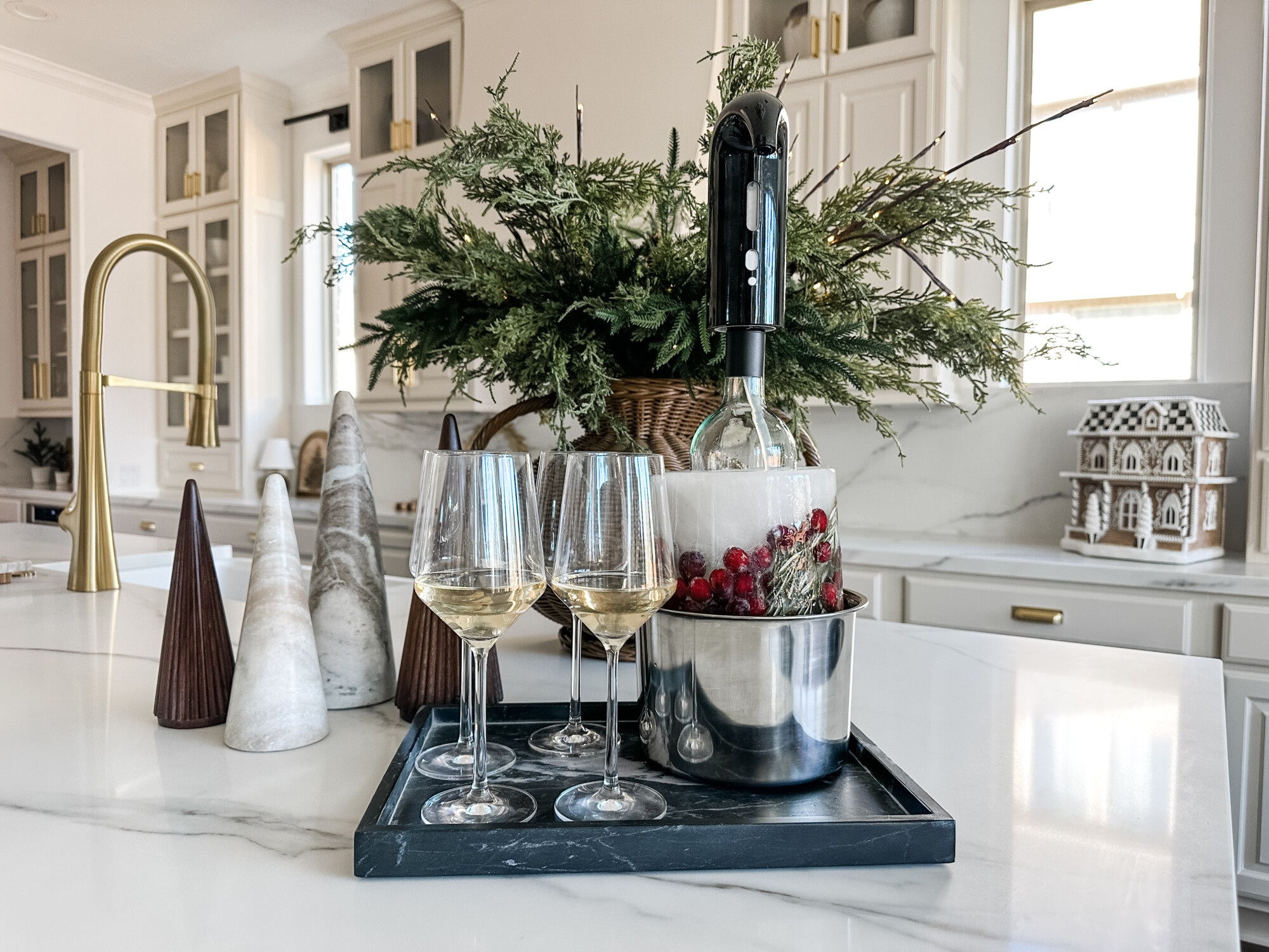Modern holiday kitchen décor with wine glasses, marble trees, and a wine chiller filled with cranberries and ice, styled with fresh greenery and a gingerbread house for a festive touch.