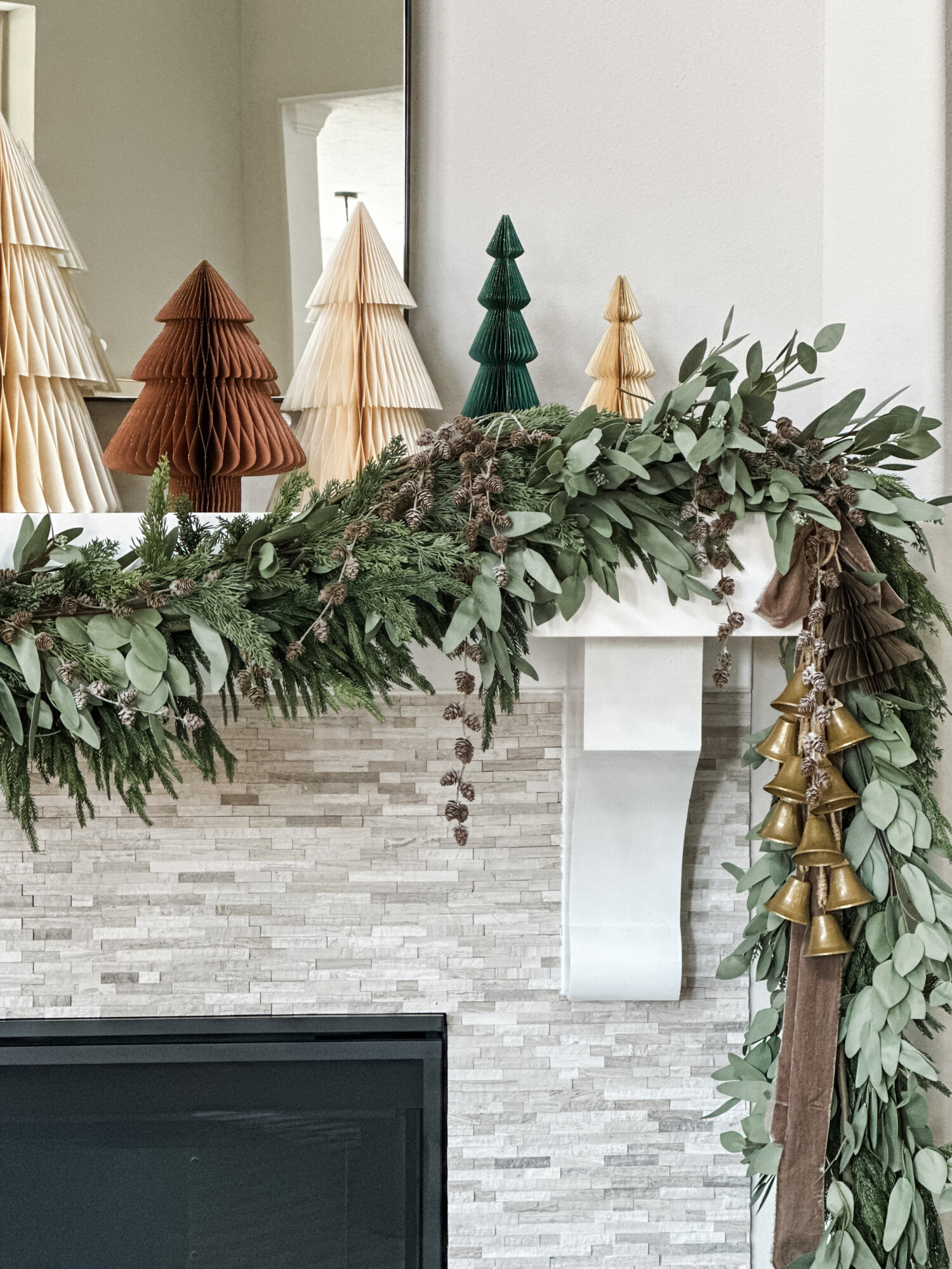 Holiday mantel decorated with fresh greenery garland, gold bells, and paper Christmas trees in neutral and green tones