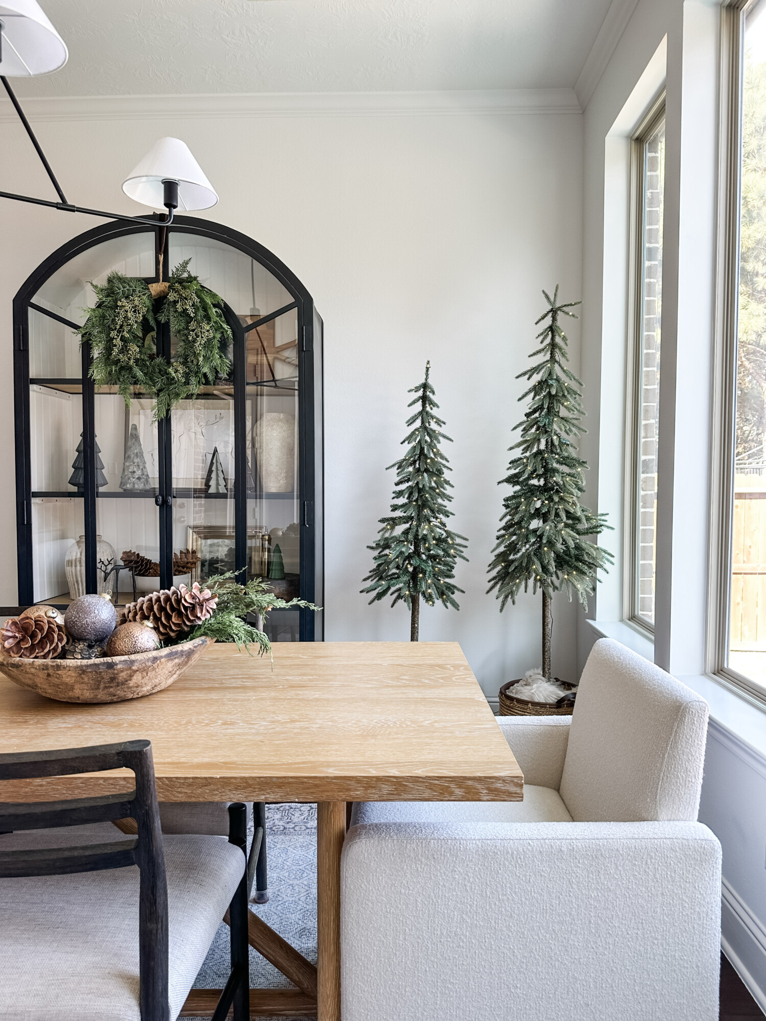 Minimal holiday dining room with slim Christmas trees, black cabinet with greenery wreath, and natural wood table décor