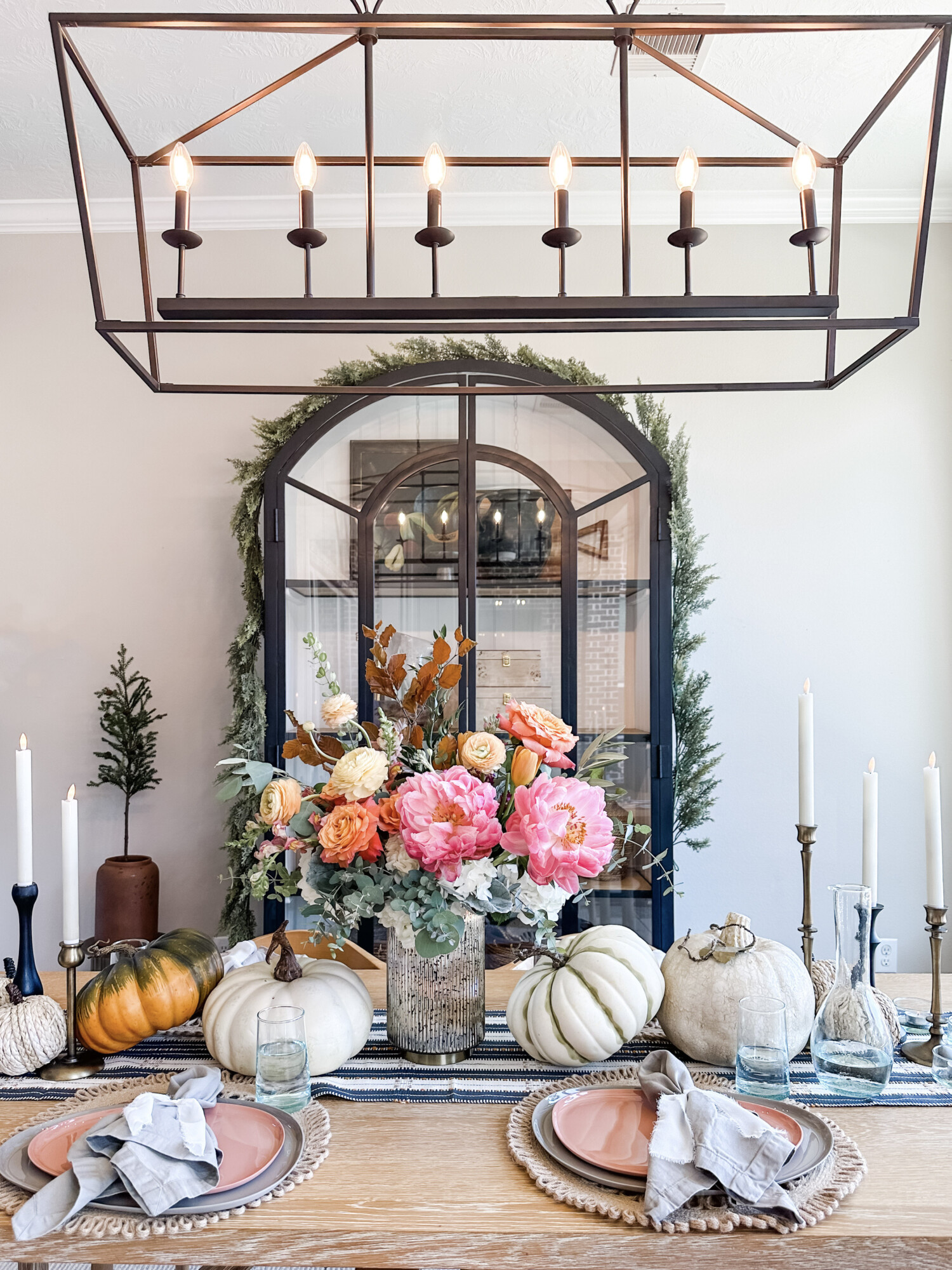 Beautiful holiday dining room setup with colorful floral centerpiece, white and orange pumpkins, striped runner, and tall candle holders under a black chandelier, ideal for fall or Thanksgiving hosting.