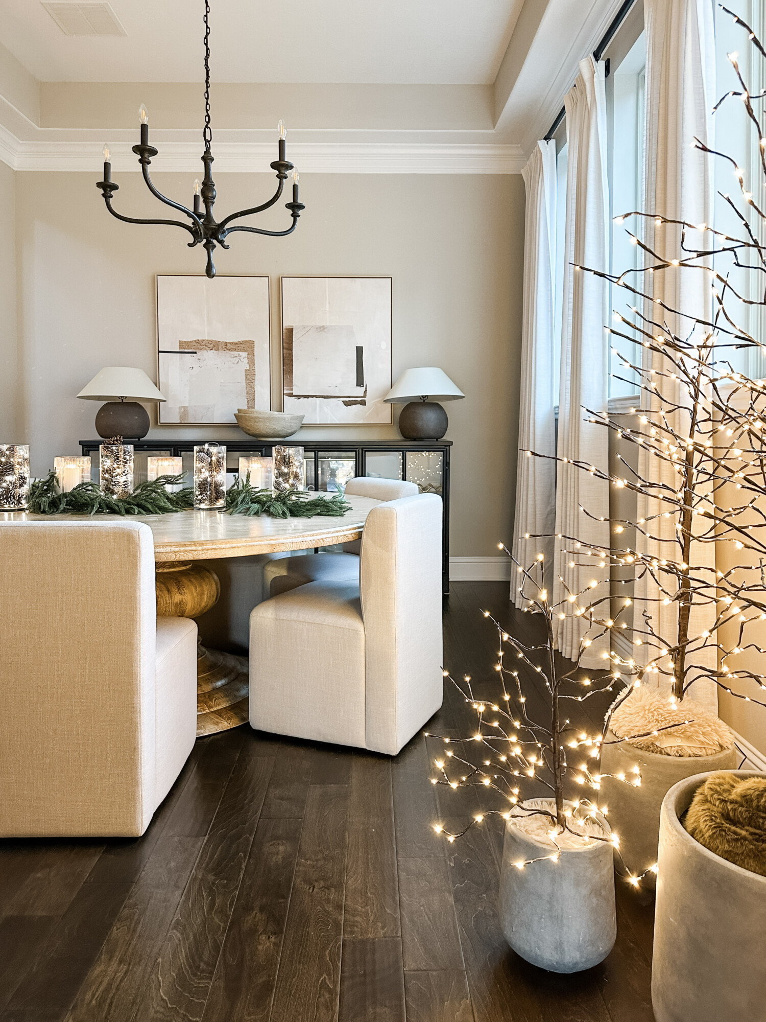 Neutral dining room styled for the holidays with a round wood table, light-up trees, greenery garland, and soft ambient lighting for a cozy Christmas home atmosphere.