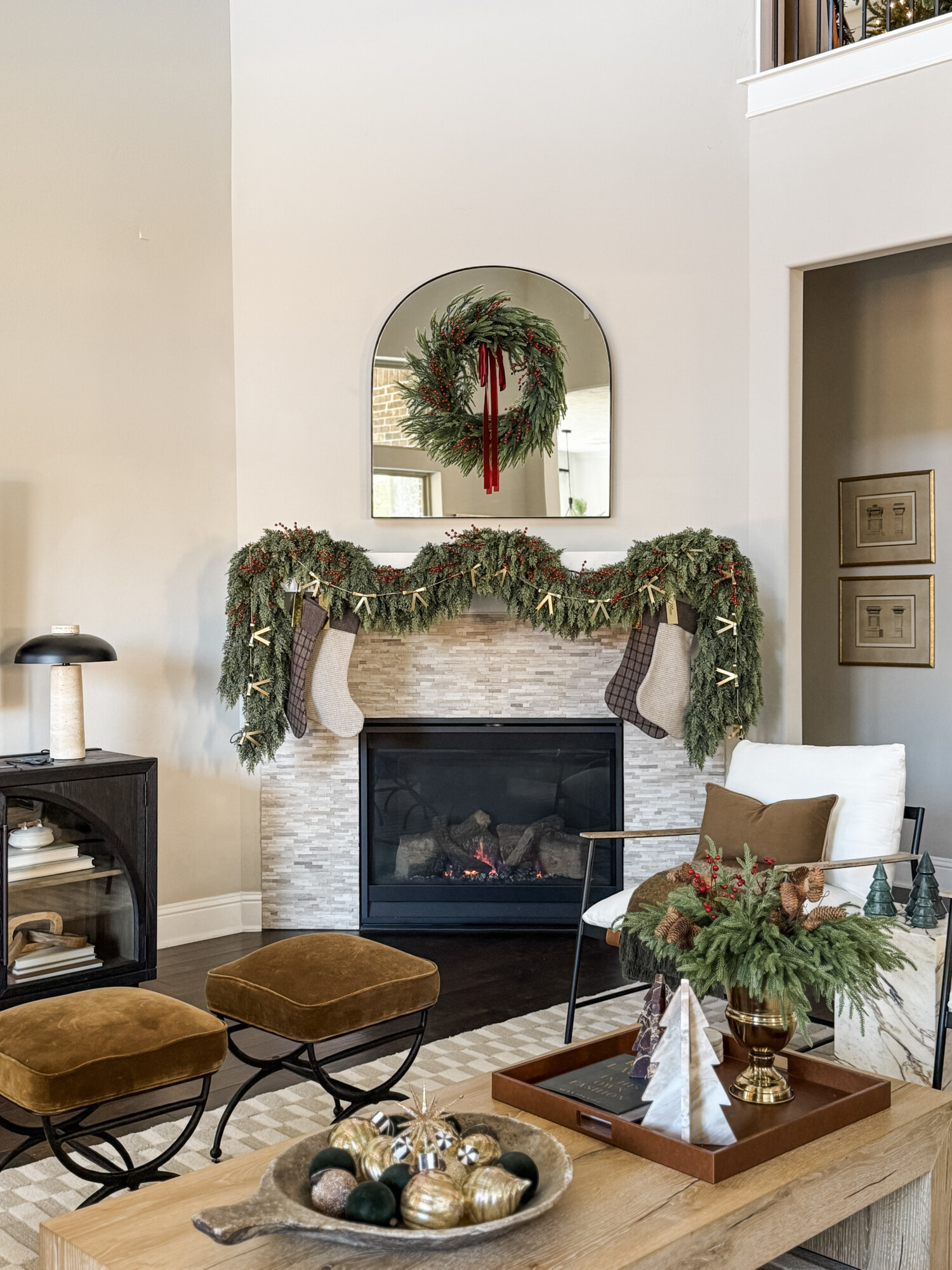 Living room fireplace decorated with lush holiday garland, a wreath with velvet ribbon, neutral stockings, and warm seasonal accents styled around a soft neutral palette.