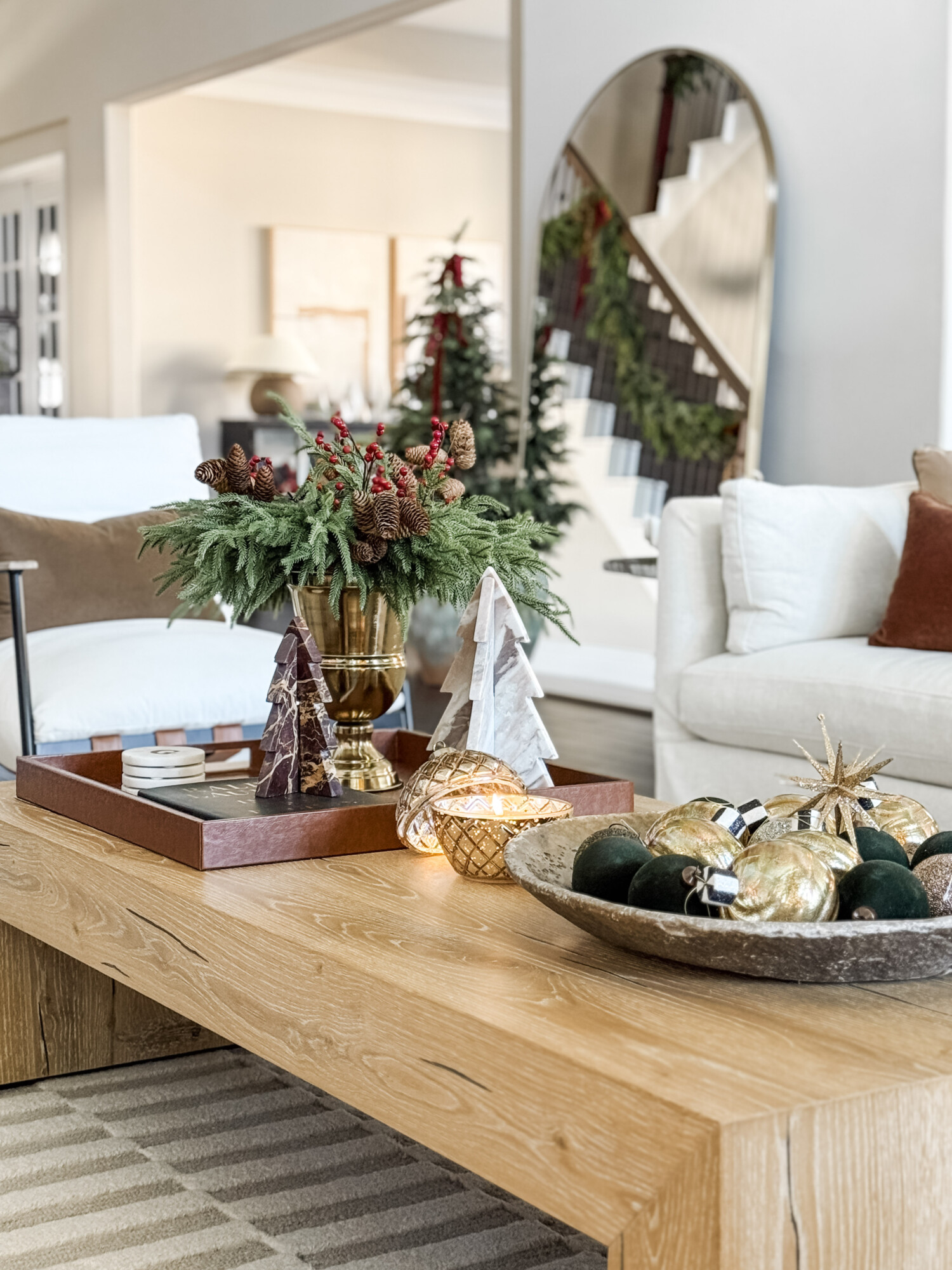 Coffee table styled with greenery in a gold vase, sculptural trees, gold and green ornaments, and warm candlelight for an elevated holiday display.