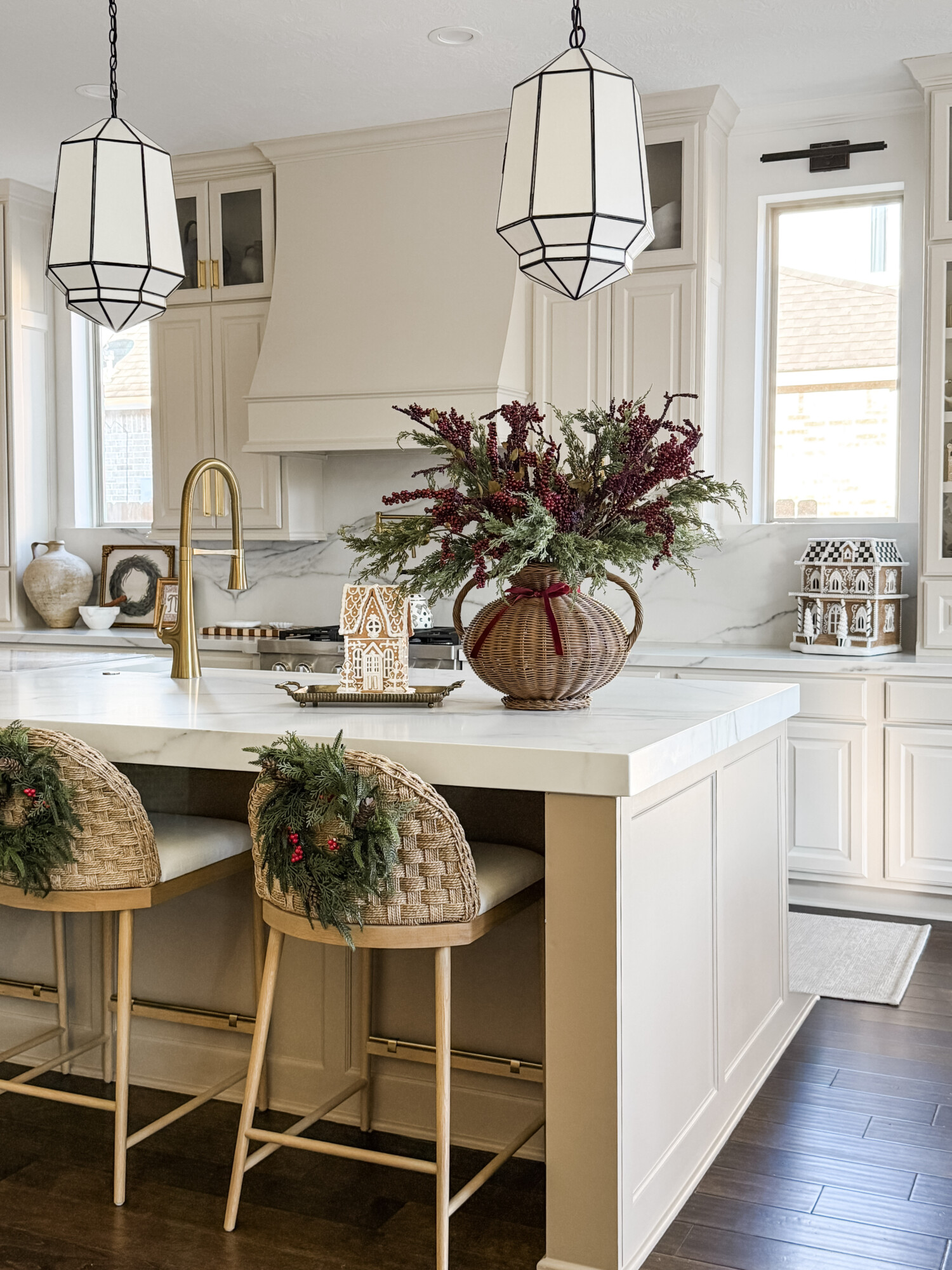 Bright kitchen with barstools decorated with small wreaths, a woven vase filled with holiday greenery, gingerbread houses, and soft seasonal accents on a white countertop.