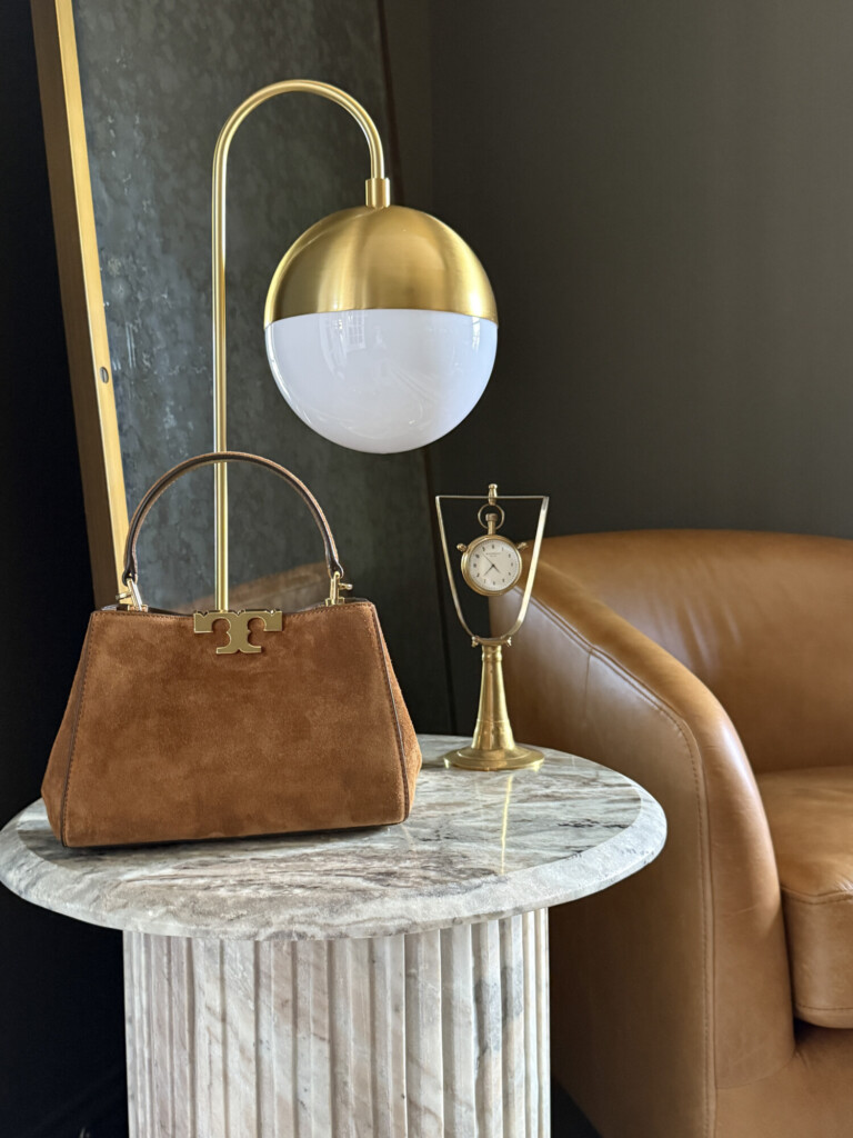 Brown suede handbag styled on a marble side table with a brass lamp and leather chair
