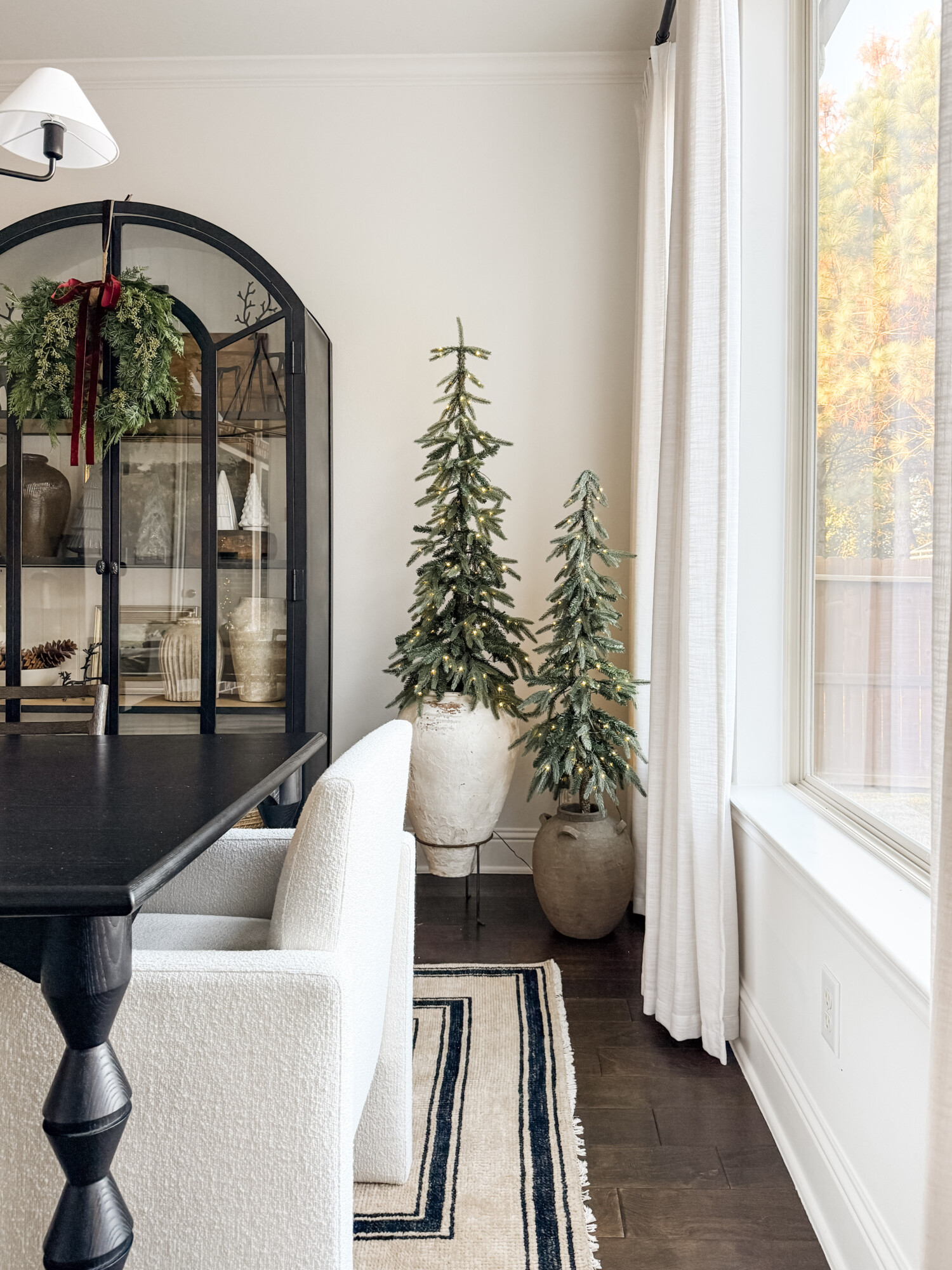Dining room corner with two slender Christmas trees in stone planters, a black display cabinet with a holiday wreath, and soft neutral curtains creating a subtle seasonal moment.