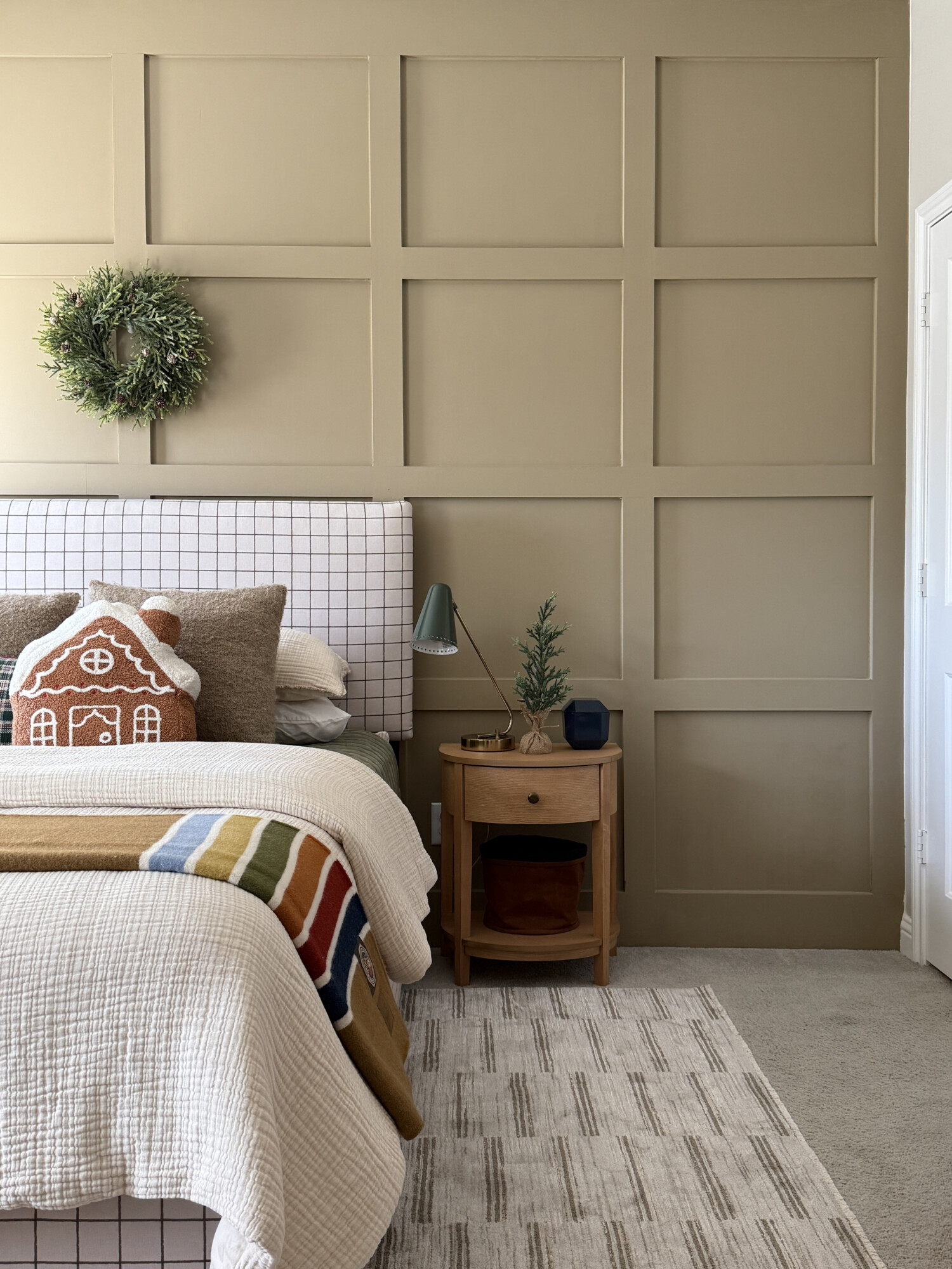Bedroom featuring chambray bedding, a gingerbread pillow, a textured throw, a small tabletop tree, and a neutral paneled wall for a soft Christmas look.