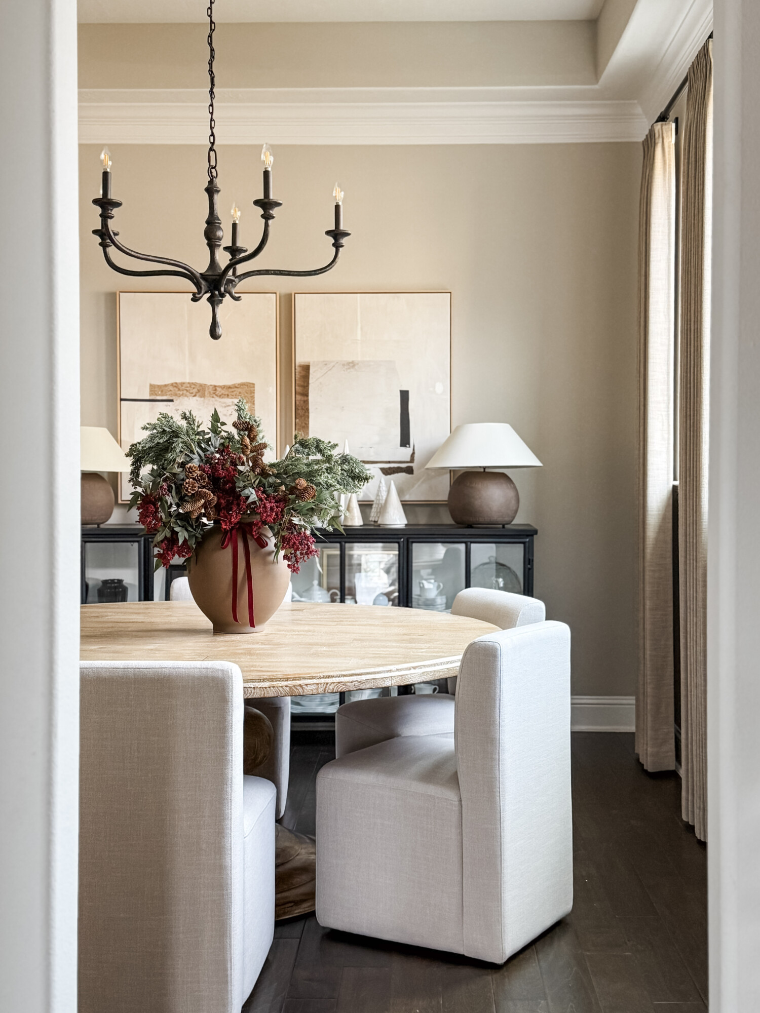 Dining room with a round table, upholstered chairs, a terracotta vase filled with holiday greenery and berry stems, and neutral artwork creating a warm and elevated seasonal moment.